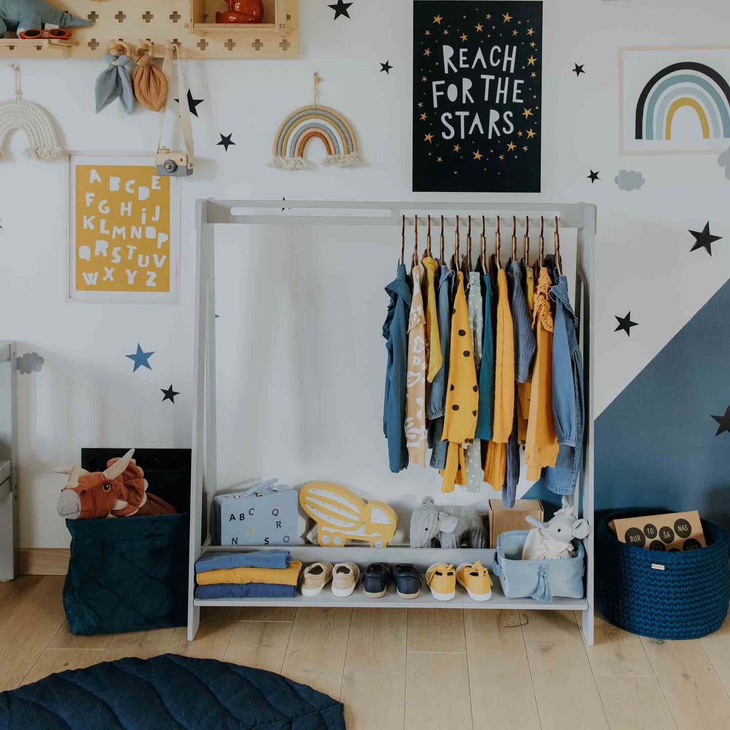 A child's closet beautifully organized with clothes hanging from a kids' clothing rack, a shelf neatly arranged with shoes and toys, and whimsical wall decorations such as stars, rainbows, and a "Reach for the Stars" poster. This enchanting space also includes a wooden kids' clothing rack that is perfect for dress-up storage.