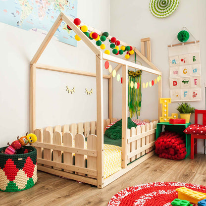 A child's room featuring a wooden platform house bed with a picket fence, colorful garlands, alphabet wall decor, a map, and various toys. The space is adorned with bright, cheerful colors and cozy knitted items.