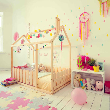 Child's bedroom featuring a platform house bed with a picket fence, colorful wall decorations, stuffed animals, a pink balloon, and a white crate with more toys. The room has a playful and cozy atmosphere.