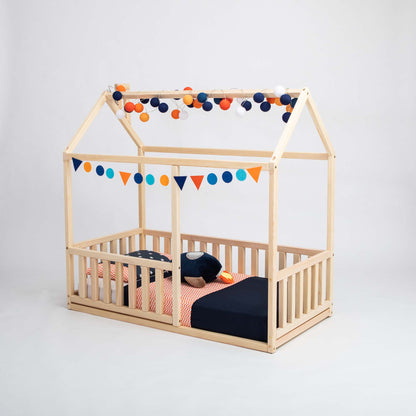 Montessori floor house bed with rails for children, featuring a vertical rail fence design, decorated with a triangular flag banner and string of round lights. The preschool bed is made up with a navy blue comforter, orange accent pillows, and red-patterned sheets.