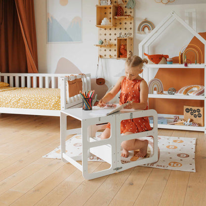 A young girl in an orange dress is sitting at a white desk, drawing with crayons in her neatly organized room, which features a bed, shelves, and various decorative items. Among them is her favorite 2-in-1 Transformable Kitchen Tower - Table and Chair Set that she uses to become the young chef of her dreams.