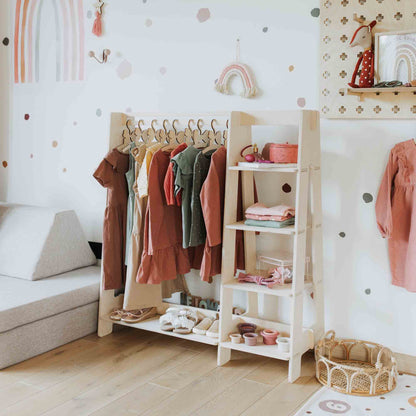 A Montessori-inspired children's room features a Children's wardrobe, dress up clothing rack in white showcasing colorful dresses, storage shelves with toys and folded clothes, and a wall adorned with rainbow decorations. A gray sofa is partially visible on the left.