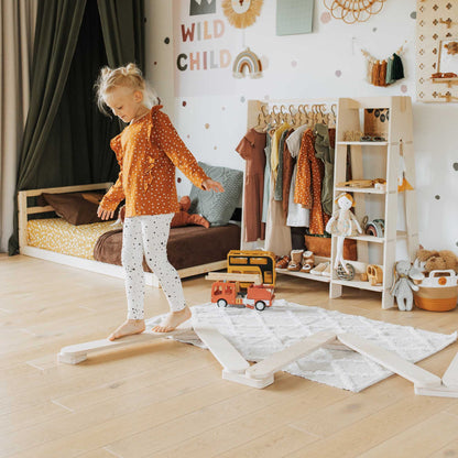 A little girl is standing in a room full of toys, including a Sweet Home From Wood Balance Beam Set and climbing arch.