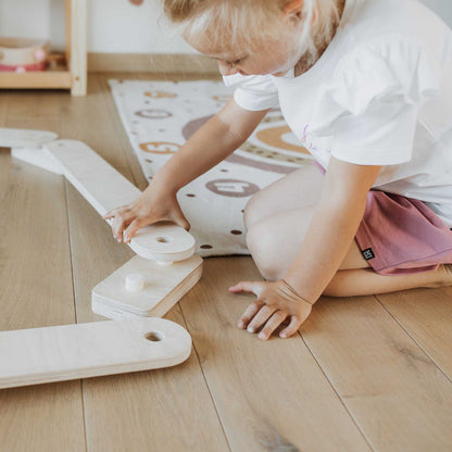 A little girl playing with Sweet Home From Wood balance beam set and wooden blocks on the floor.