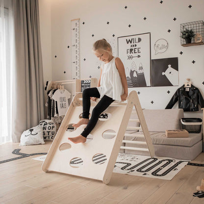 A little girl is standing on a Transformable climbing triangle + Foldable climbing triangle + a ramp in a room.