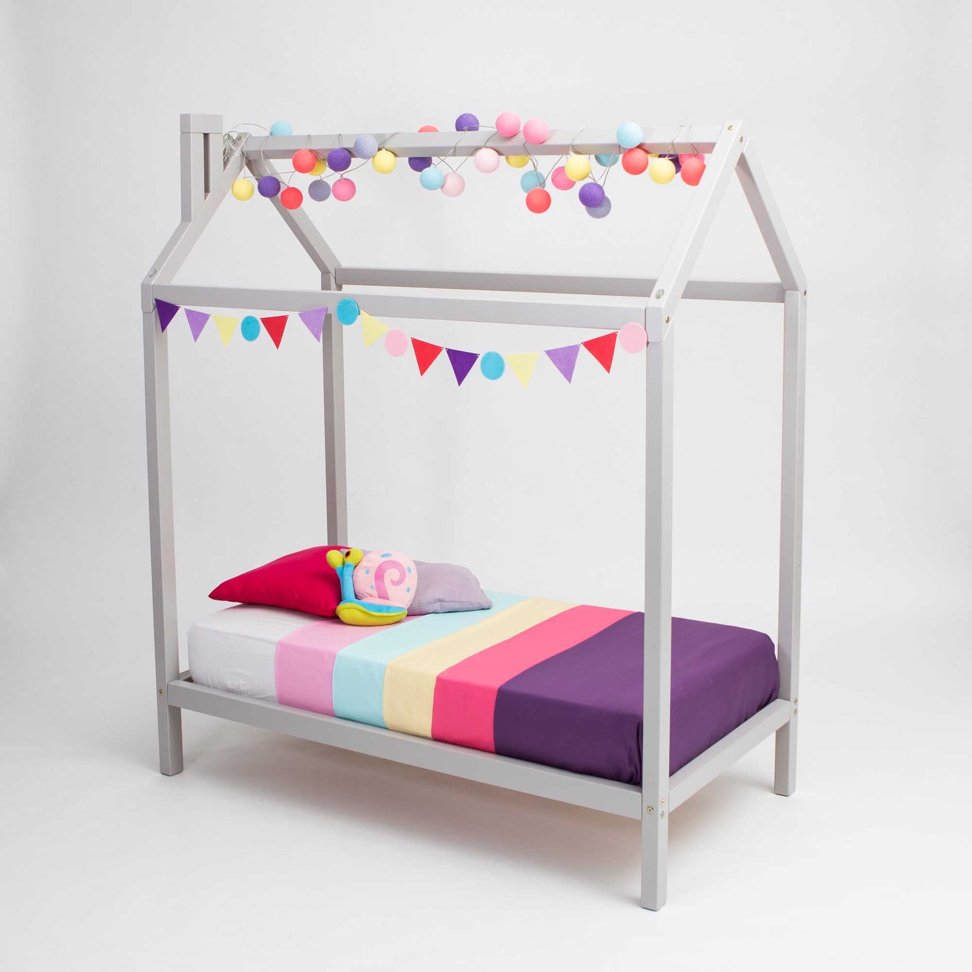 A wooden house bed on legs with pom poms and a colorful bed sheet.