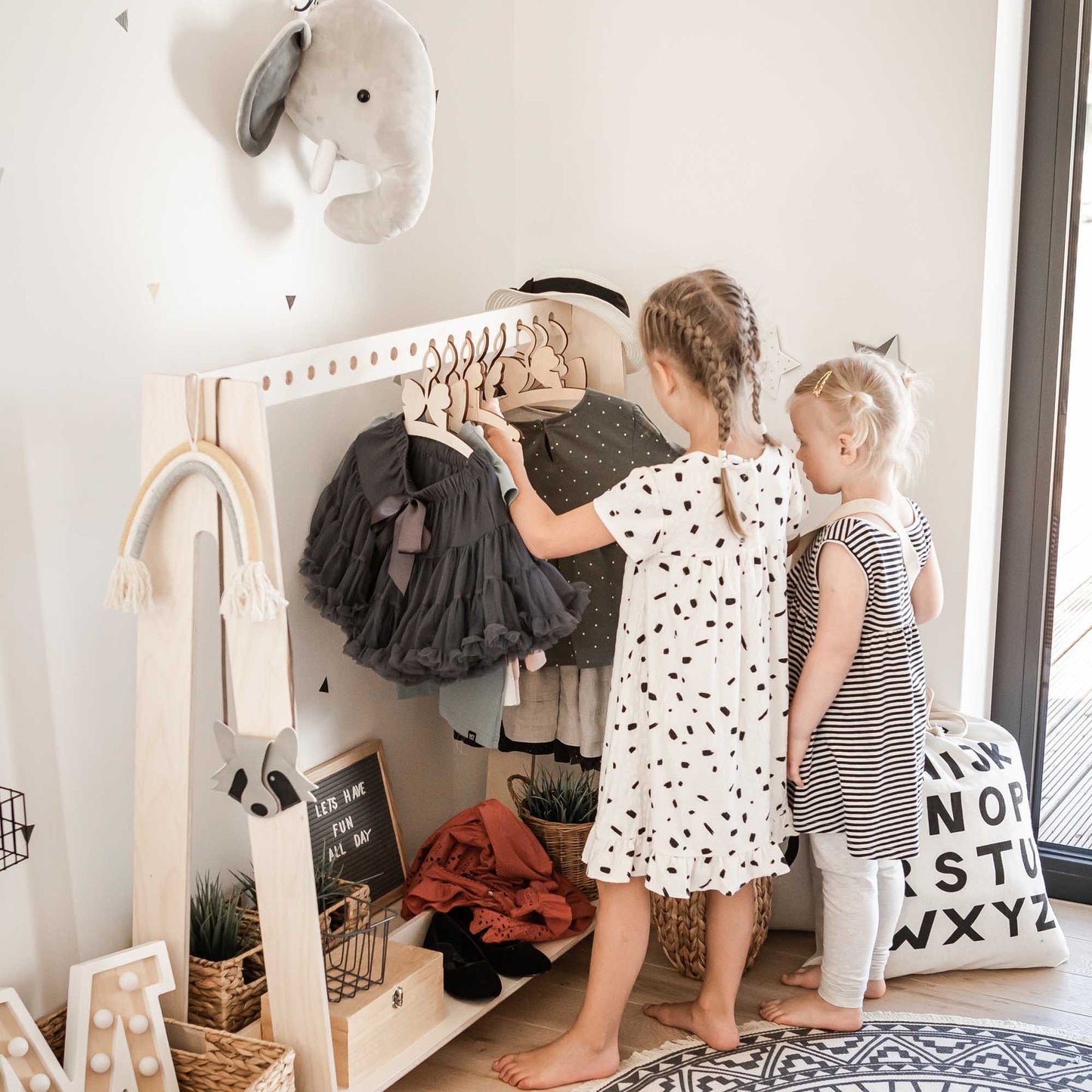 Two little girls exploring a Sweet Home From Wood A-frame kids' clothing rack filled with a shop display of clothes.