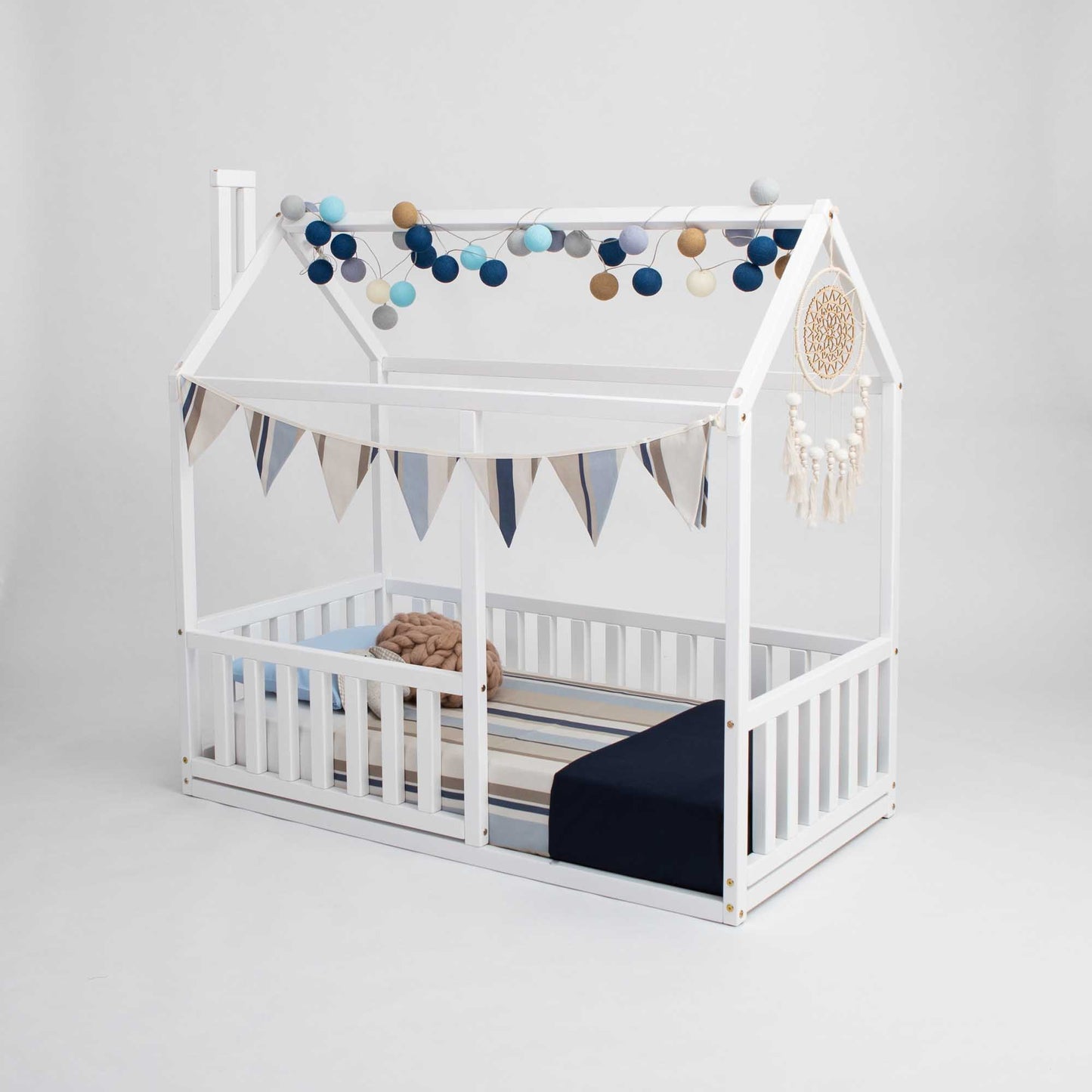 A cozy sleep haven with a Sweet Home From Wood Montessori floor house bed with rails featuring a white baby bed with a blue canopy and bunting.