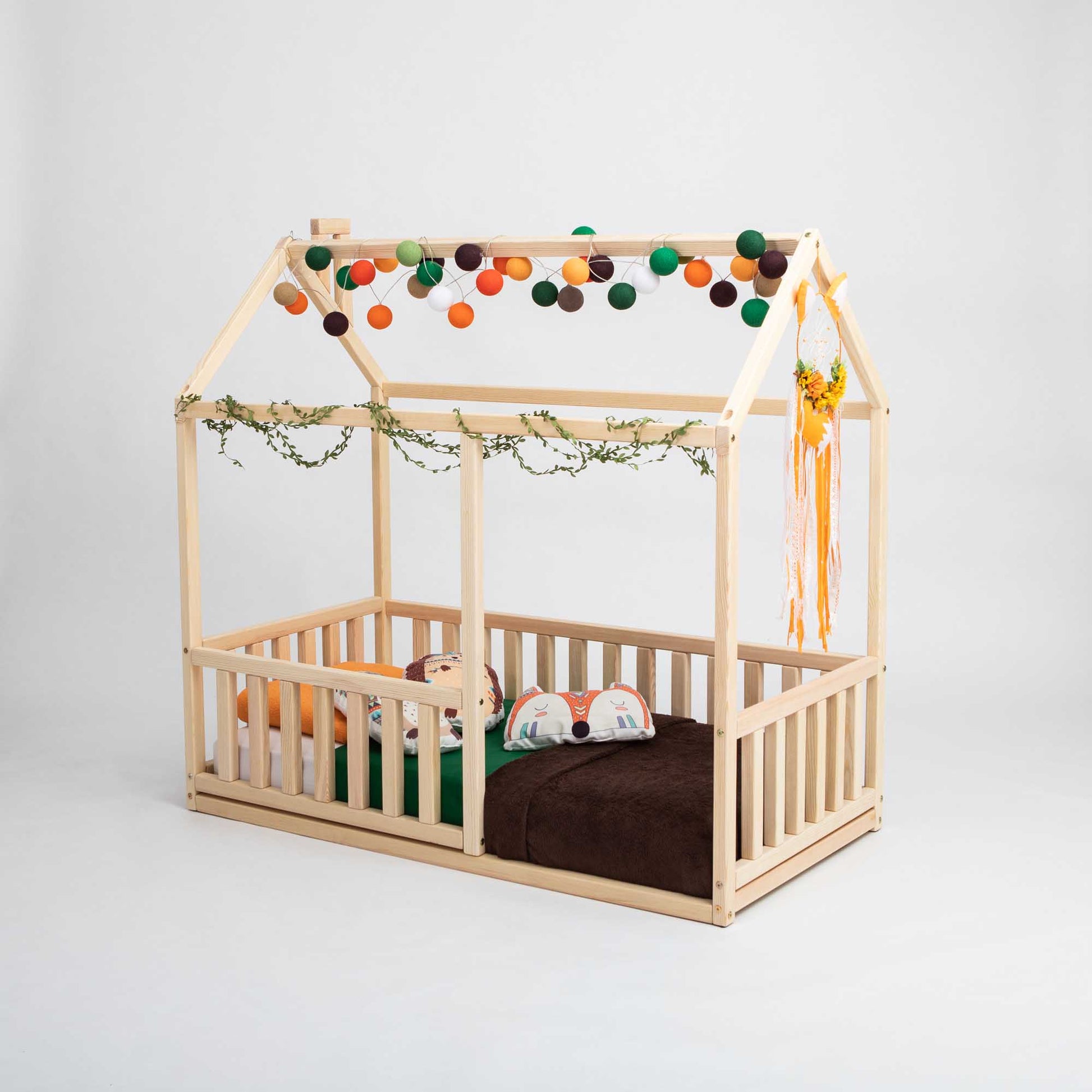 A cozy sleep haven for children, this Sweet Home From Wood Montessori floor house bed with rails features wooden construction with vertical rail fences and charming bunting and pom poms.
