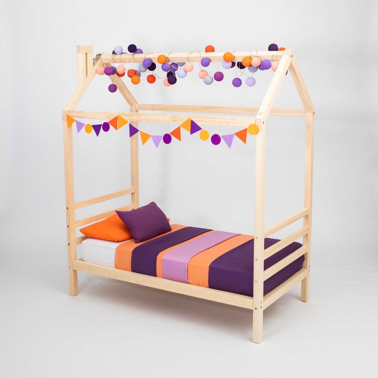 A Kids' house bed on legs with a headboard and footboard with purple and orange pom poms hanging from the ceiling.
