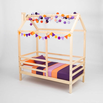 A Wooden house bed on legs with a fence with purple and orange pom poms.
