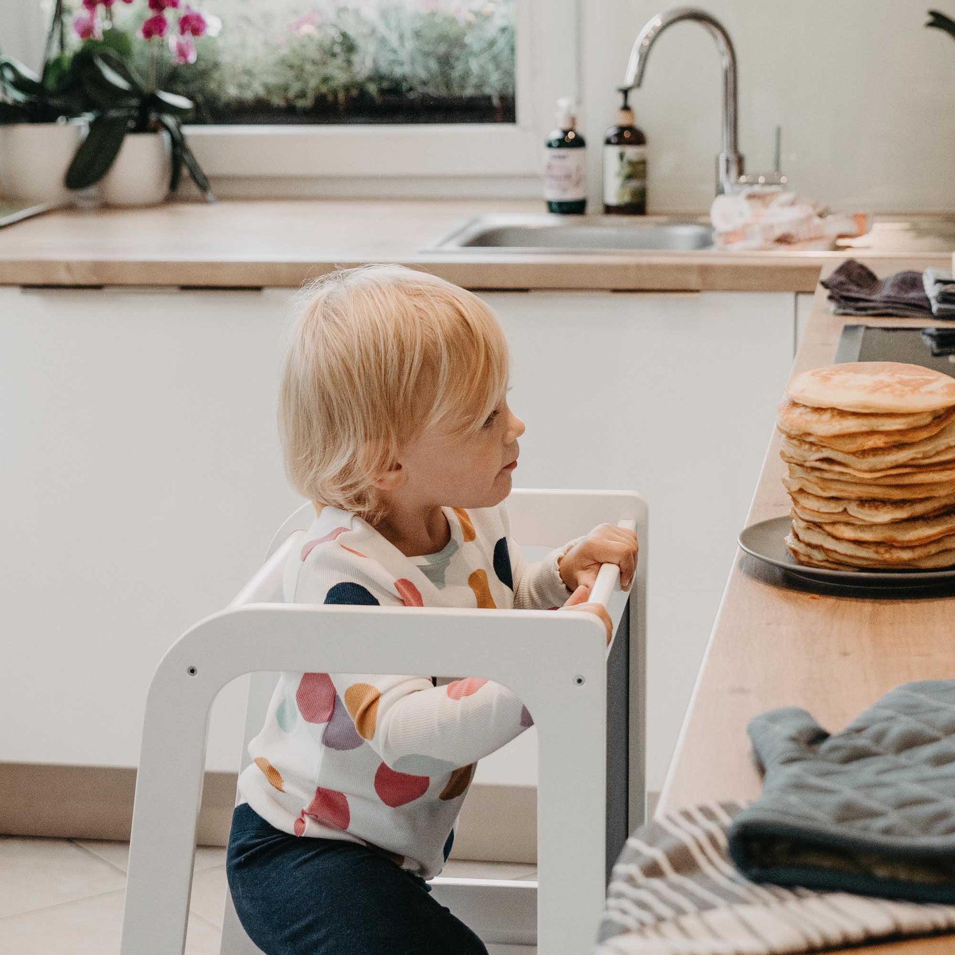 A toddler sitting on a Sweet Home From Wood kitchen tower with blackboard in the kitchen using a kids step stool.