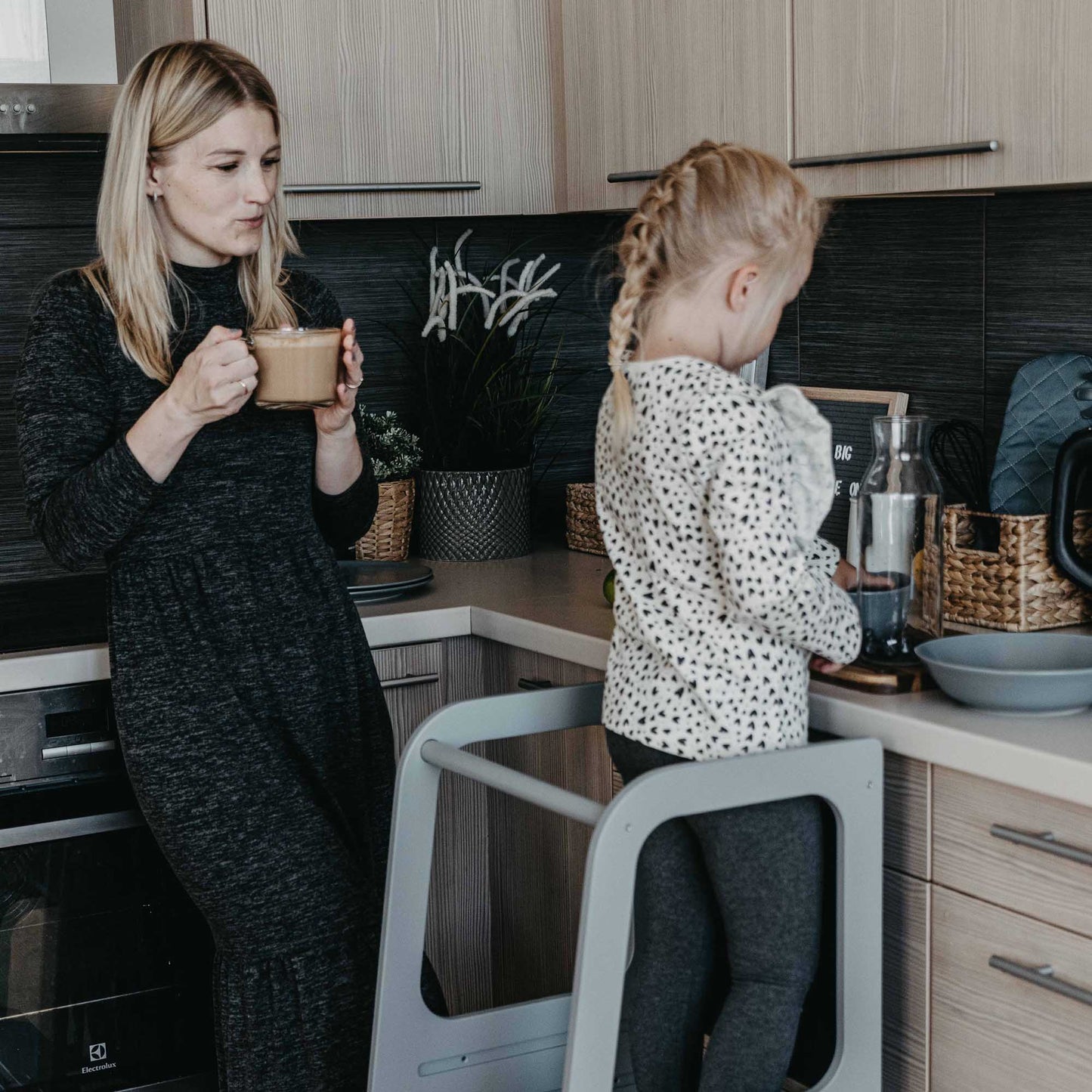 Girl cooking on a toddler step stool while mother drinks her coffee.