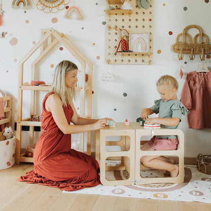 A woman and a child sitting at a wooden table in a playroom, using the Sweet Home From Wood 2-in-1 transformable kitchen tower - table and chair set.