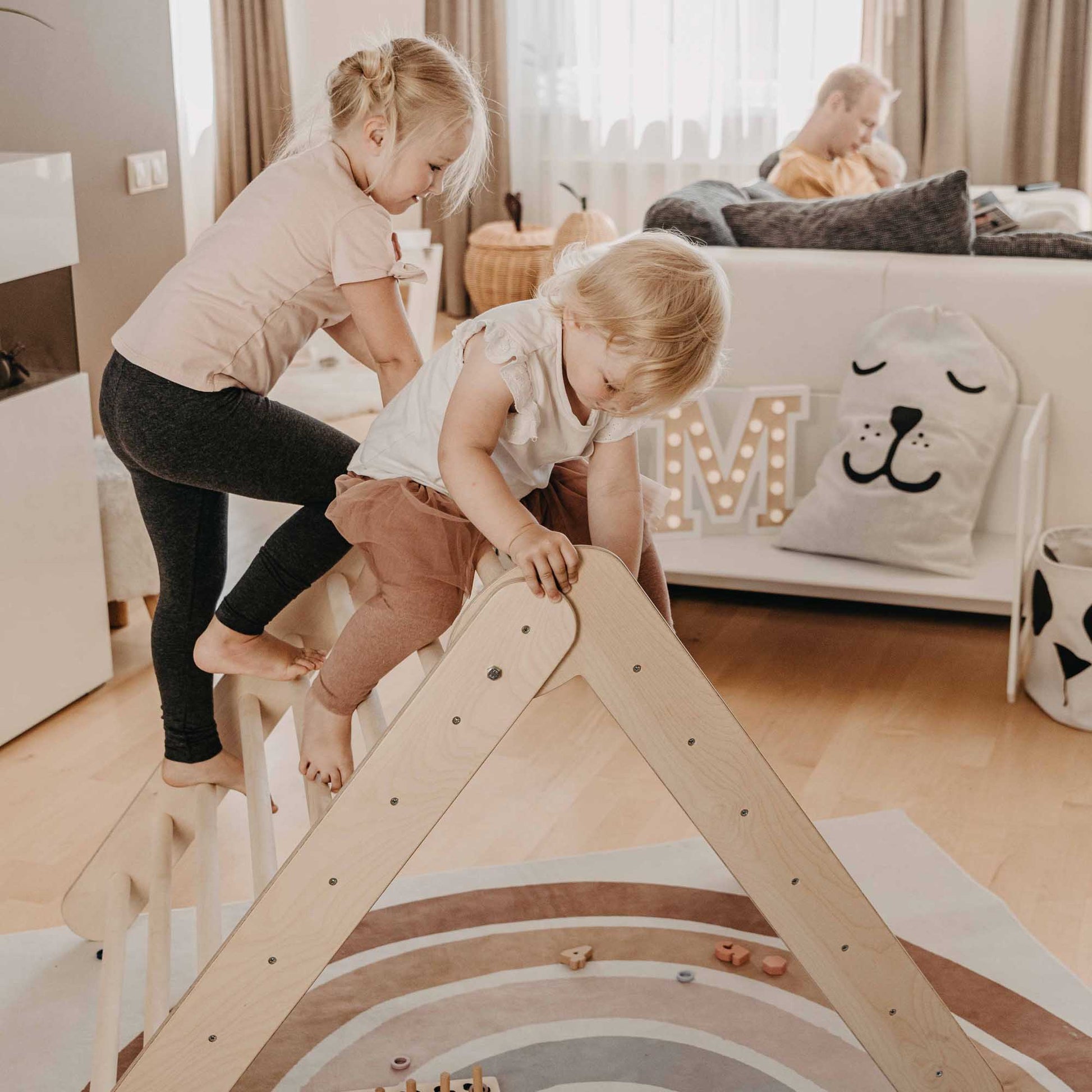 Two children, while playing with a foldable climbing triangle with 2 slope levels in a living room, are enhancing their motor skills.