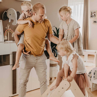 A man is playing with his children on Montessori furniture in a living room, helping them develop their motor skills with the Foldable climbing triangle with 2 slope levels.
