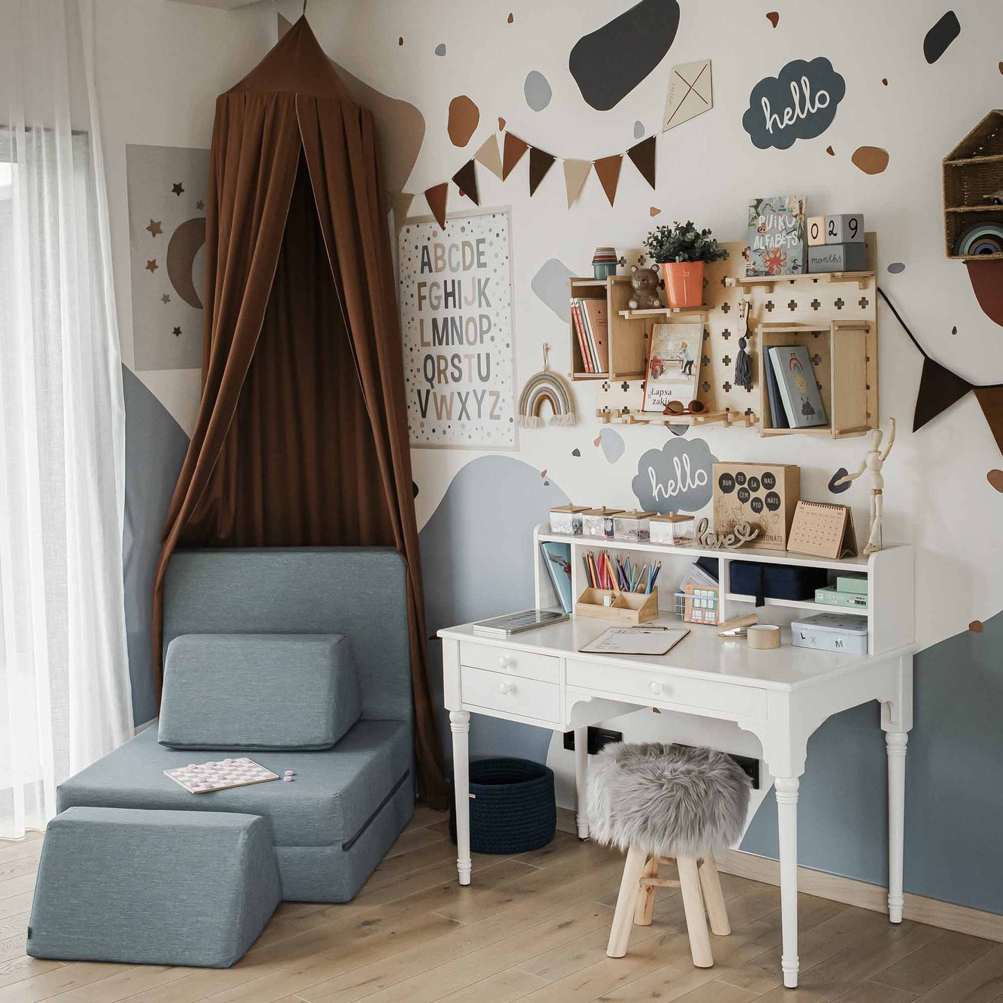 A children's playroom features a reading nook with a brown canopy, a versatile pedestal desk with organized stationery and adjustable leg height, wall decorations, alphabet art, and a table with a hutch filled with toys and books.