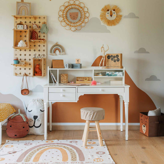 A children's room with a versatile table with a hutch, a wooden stool, wall shelves for optimal storage, and decor items including rainbows and animal-themed toys. The walls feature cloud decals and an orange mural. Effective cable management ensures the space stays tidy.