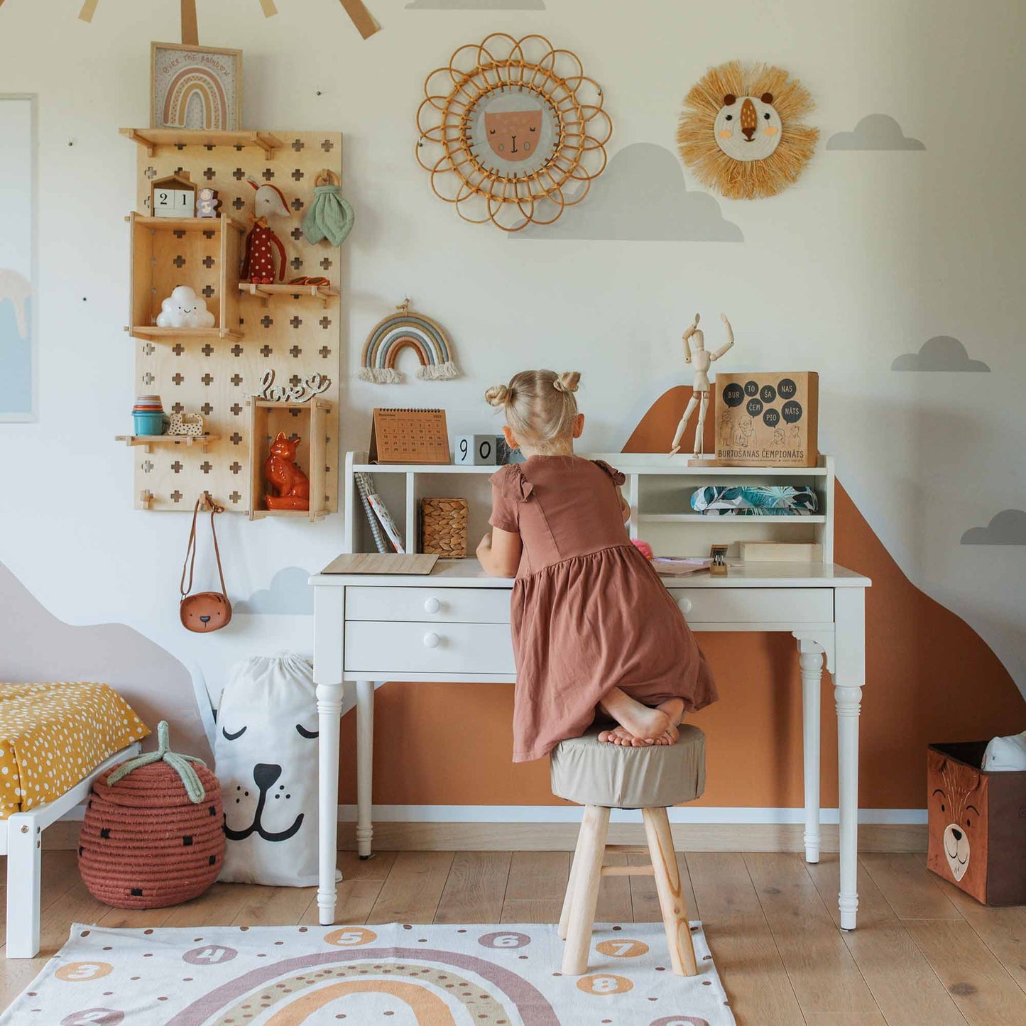 A young child stands on a stool, reaching for objects on a versatile Table with a Hutch in a decorated room with shelves, a wall clock, and various items like bags, toys, and rainbow-themed decor. The desk's cable management and storage options keep everything neat and organized.
