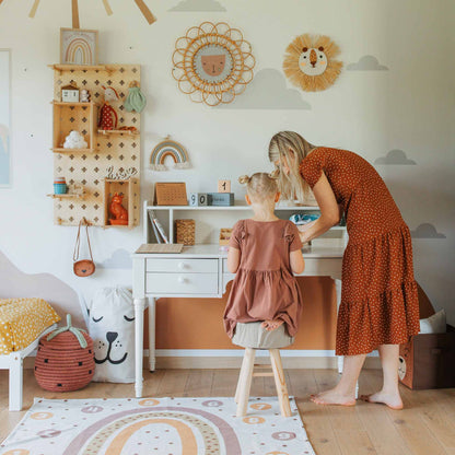 A woman and a child are in a playroom adorned with animal-themed wall art, wooden toys, and a white pedestal desk. The woman is leaning over the desk, engaging with the child seated on a stool. A table with a hutch in the corner provides ample space for organizing their things.