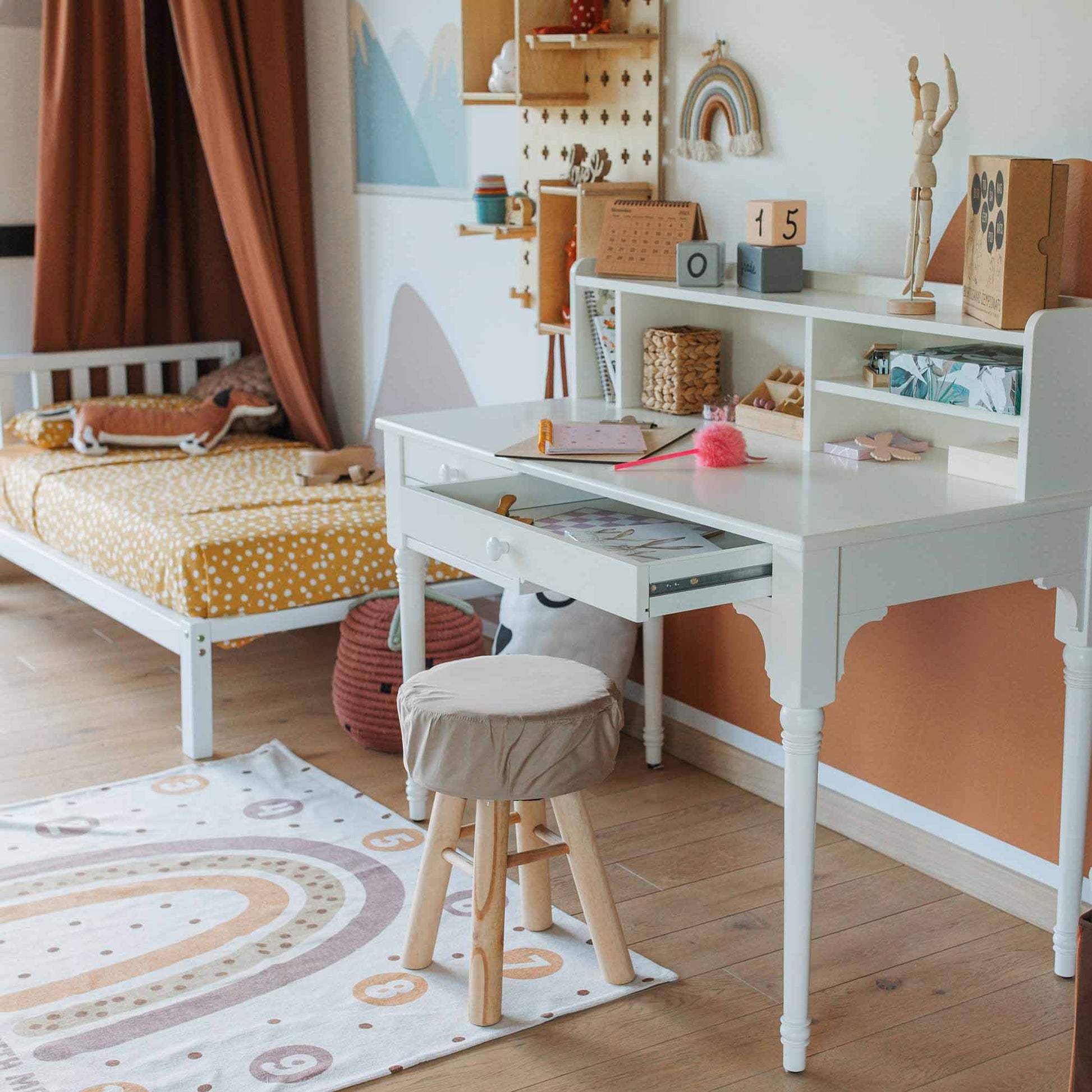 A children's study area features a versatile table with a hutch and a pedestal desk, accompanied by a stool and various items. Adjacent to the desk is a bed adorned with a polka-dotted yellow blanket. The room boasts wooden flooring, a decorative rug, and ample cable management for tidy organization.