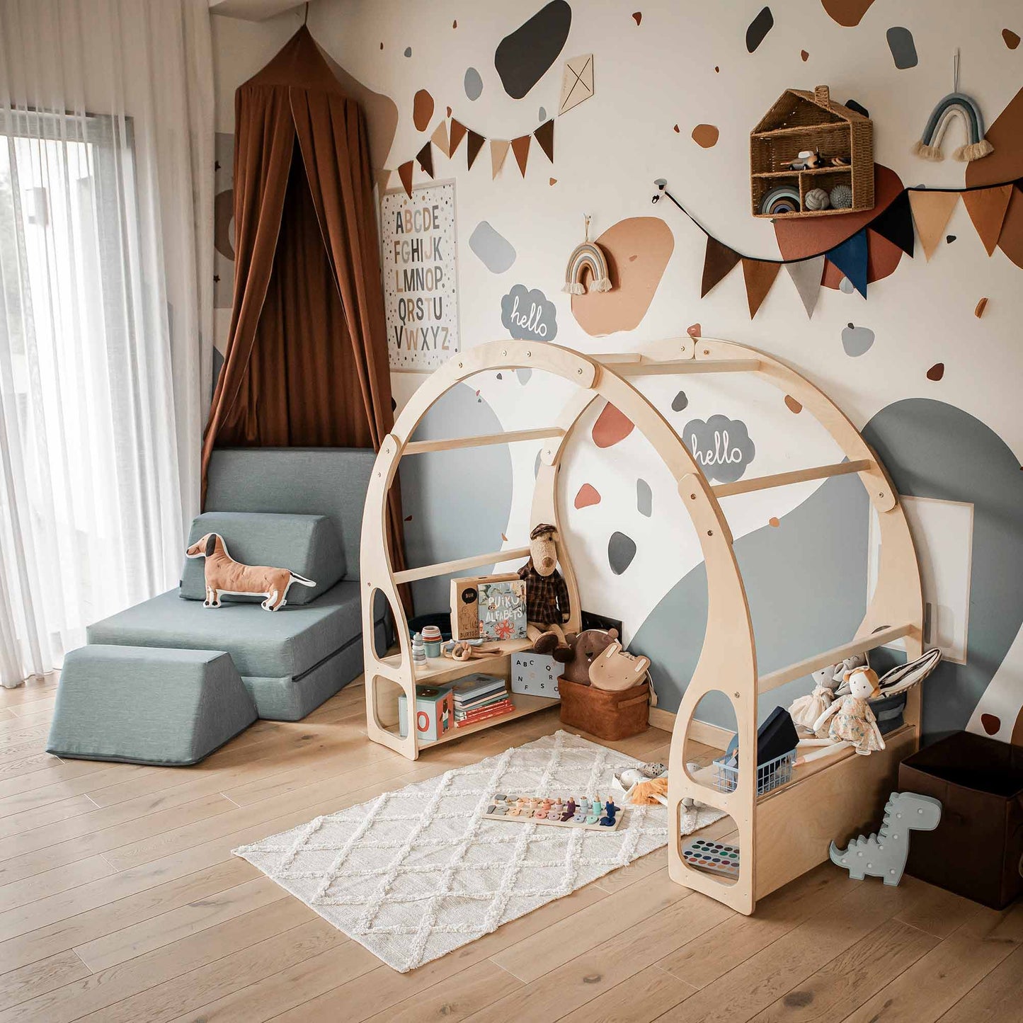 A modern children's room with a wooden tent-like bed, a play area featuring a Play stand for toddlers designed for imaginative play, a blue armchair, and alphabet decor on the wall. Light wood flooring and a white and pastel abstract mural complete the space, fostering child development effortlessly.