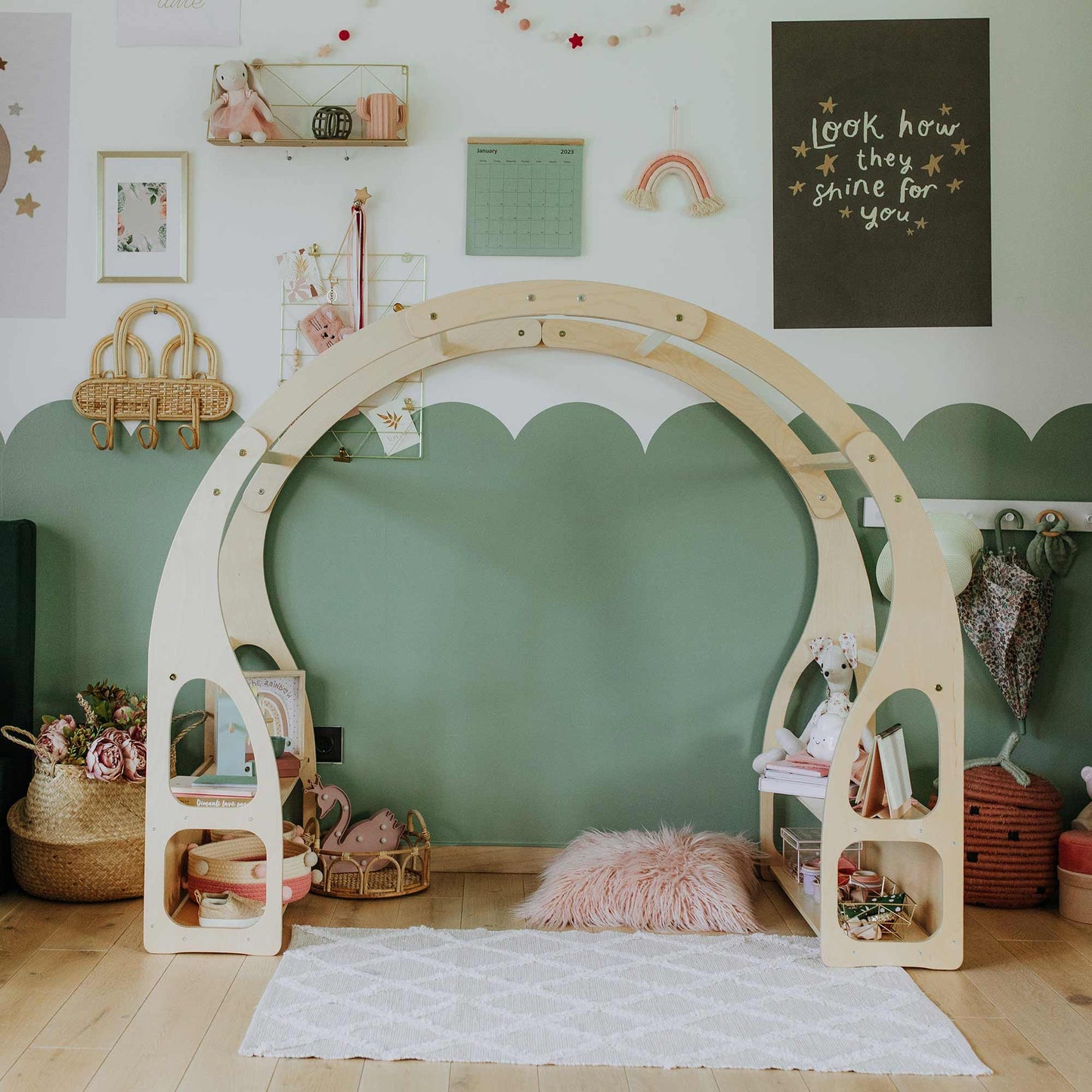 Children's playroom with a wooden arch structure, various Montessori toys, baskets, wall art, and a pink fluffy cushion on a white patterned rug. The wall is painted with a green and white scalloped design to nurture child development. A versatile Play stand for toddlers keeps the space organized and engaging.