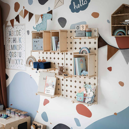 A neatly organized nursery room features wall shelves with books, toys, and decor items. The walls are decorated with alphabet prints, buntings, and a blue "hello" sign. A versatile Large Pegboard Floating Display Shelf from Sweet Home from Wood adds a practical storage solution to keep everything tidy.