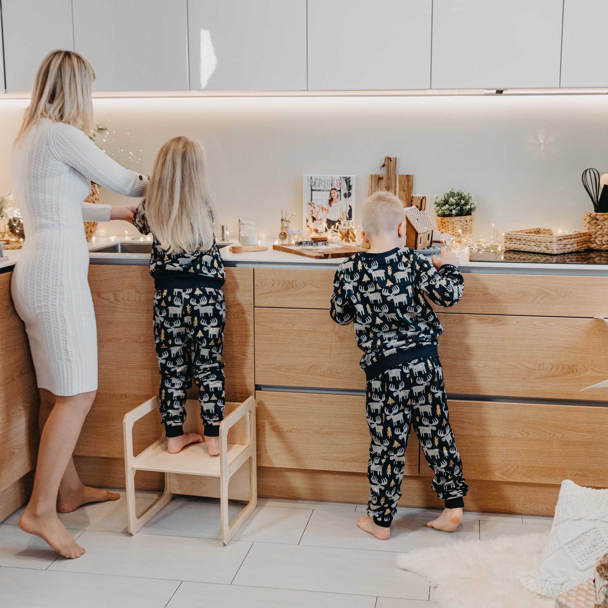 A woman and two children, dressed in pajamas, sitting at a Sweet Home From Wood Montessori weaning table and chair set in their kitchen.