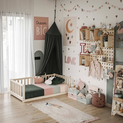 A cozy child's bedroom featuring a low-profile Montessori bed with three-sided rails, a canopy, and wooden storage shelves. The walls are decorated with heart patterns, art, and soft colors. Toys and books are neatly arranged.