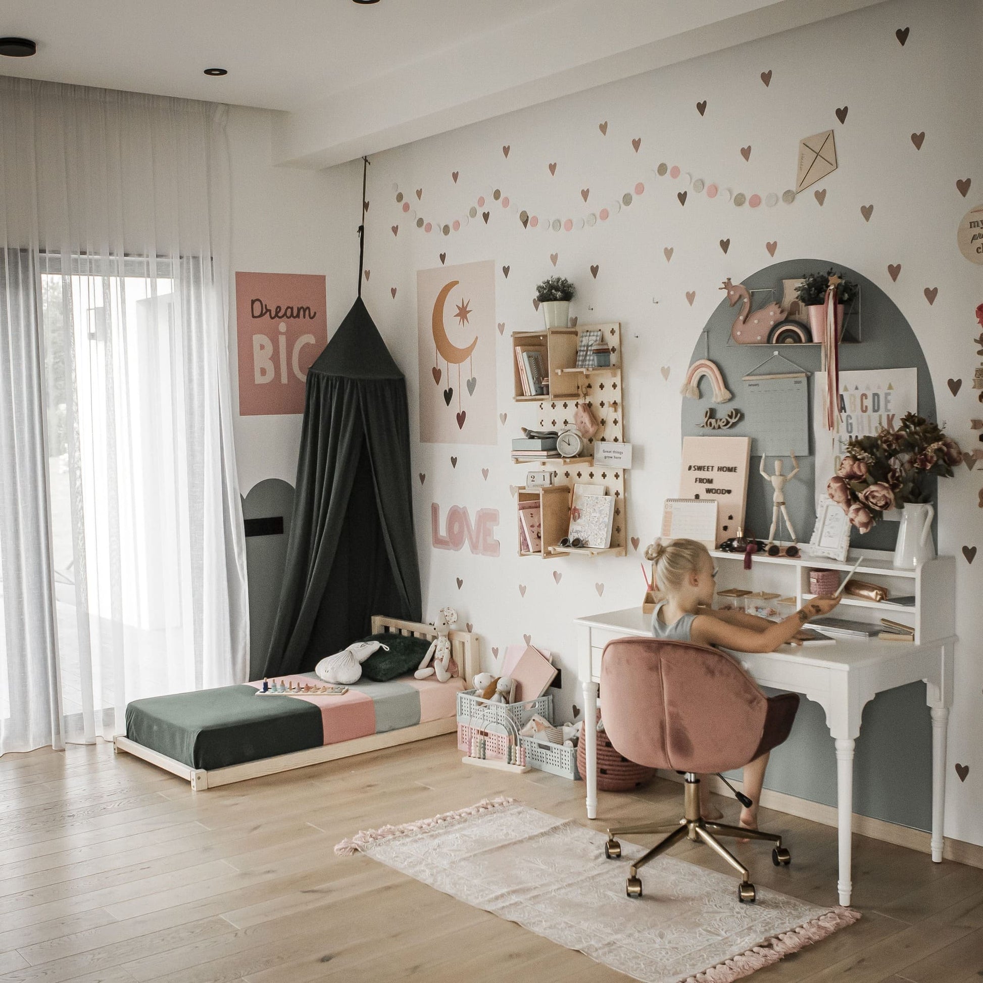 A child sits at a white desk in a decorated bedroom with heart decals on the walls, featuring a Toddler bed with a headboard and canopy inspired by Montessori principles, along with various shelves and decor items.